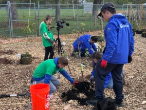 A group of young people plant a small tree in a mulched area. Some are placing soil around the tree while another holds a camera on a tripod, capturing the activity. A fence and sports fields are in the background.