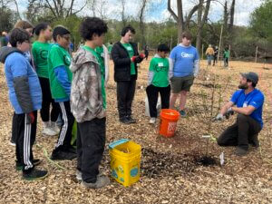 A group of kids in casual clothes stand outside on mulch, attentively listening to an adult instructor kneeling by a young tree and orange bucket, learning about planting in a wooded area.