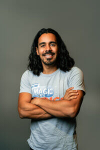 A man with long dark hair and a beard smiles with his arms crossed, wearing a light gray t-shirt that says Its not MAGIC, its SCIENCE, standing against a plain gray background, reminiscent of Tami Jo Wilt speaking at Inspire Momentum.