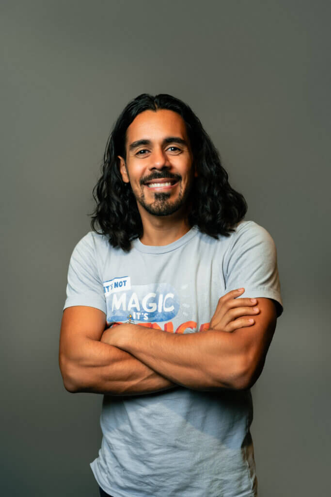 A man with long dark hair and a beard smiles with his arms crossed, wearing a light gray t-shirt that says Its not MAGIC, its SCIENCE, standing against a plain gray background, reminiscent of Tami Jo Wilt speaking at Inspire Momentum.