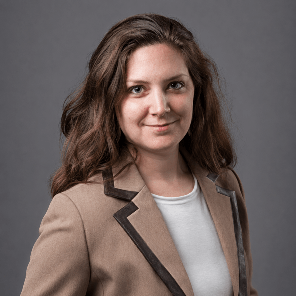A woman with long brown hair, wearing a beige blazer with dark trim over a white top, stands against a plain gray background, smiling softly at the camera—ready to attend Jason Strickland Speaking at Inspire Momentum.