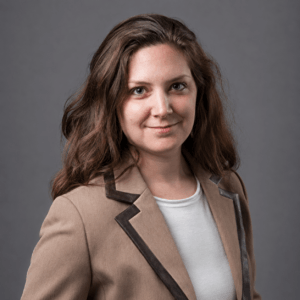 A woman with long brown hair, wearing a beige blazer with dark trim over a white top, stands against a plain gray background, smiling softly at the camera—ready to attend Jason Strickland Speaking at Inspire Momentum.