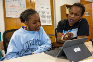 A woman helps a young girl with a tablet at a table in a classroom setting. Both are focused on the screen, with posters and shelves in the background.