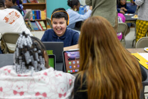 A boy wearing orange headphones smiles at a laptop while sitting with two other students in a classroom, engaging in youth tech learning and building technology career readiness through interactive STEM activities.
