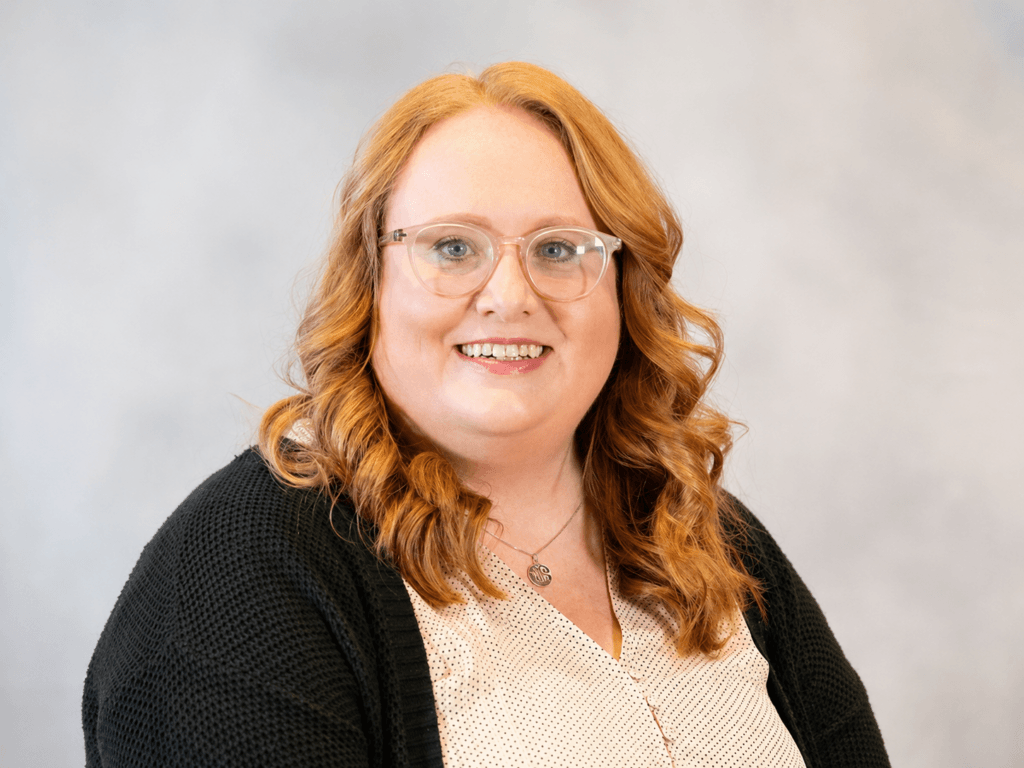 A woman with shoulder-length curly red hair and glasses smiles at the camera. She is wearing a black sweater over a light blouse and a necklace, with a neutral, blurred background at Joshua Elder Speaking at Inspire Momentum.