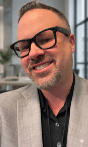 Smiling man with short hair, goatee, and black glasses wearing a light gray blazer over a black shirt, standing indoors with a blurred office background.