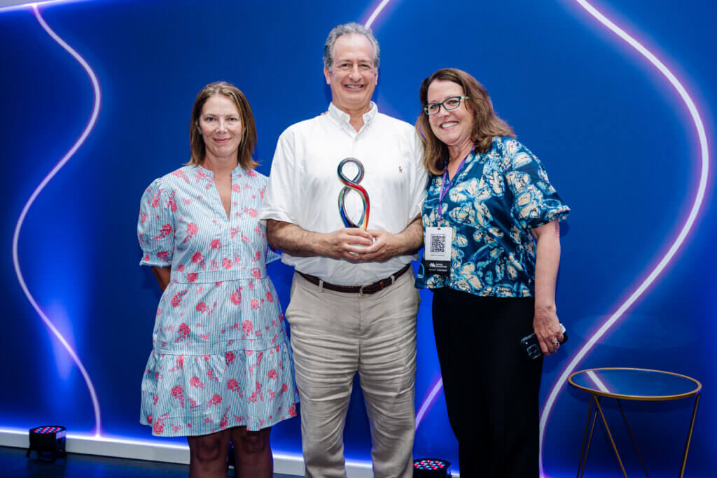 Three people stand smiling in front of a blue wall with neon lights. The man in the middle holds a colorful award, while the two women stand on either side of him. A chair is visible on the right.