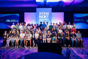 A large group of diverse young people sit and stand onstage in front of screens displaying STEM NEXT OPPORTUNITY FUND and INSPIRE MOMENTUM. The room is brightly lit with purple and blue lighting.