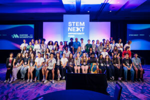 A large group of diverse young people poses together on a stage under bright lights, with screens behind them displaying STEM Next Opportunity Fund and the Inspire Momentum initiative.