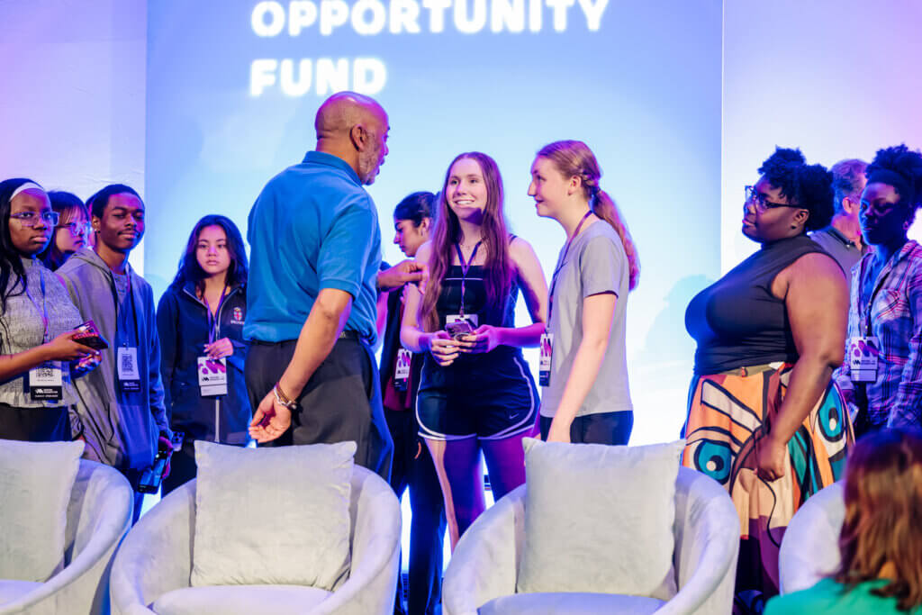 A group of people, including students and adults, stand and talk in front of a stage with the words Opportunity Fund projected on a blue-lit background. Some are wearing lanyards, as they gather to inspire momentum. A few armchairs are in the foreground.