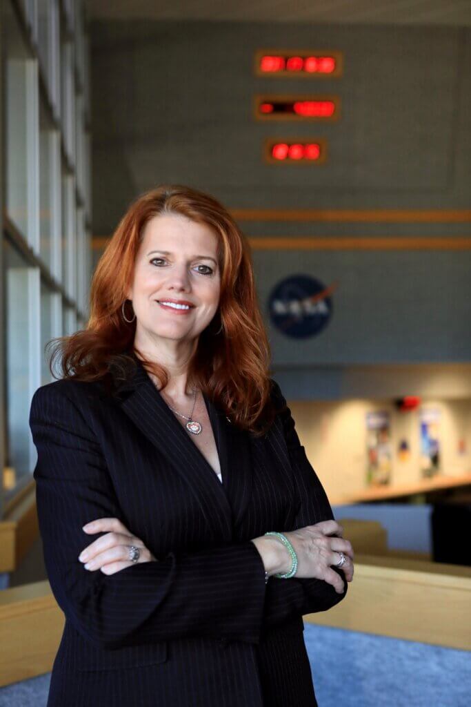 A confident woman with long red hair, wearing a dark pinstripe blazer, stands with folded arms in an indoor setting. The NASA logo and digital clocks are visible behind her, reminiscent of Jay Flores speaking at Inspire Momentum.