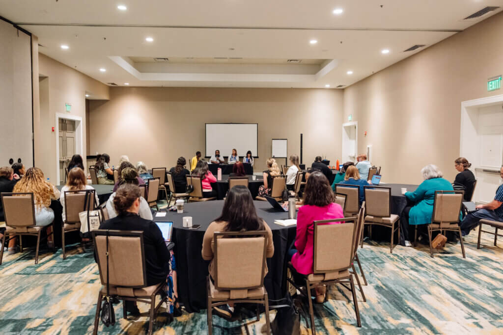 A group of people sit at tables in a conference room, facing a panel of five speakers at the front. The room has neutral walls, a projector screen, and attendees appear engaged in listening or taking notes.