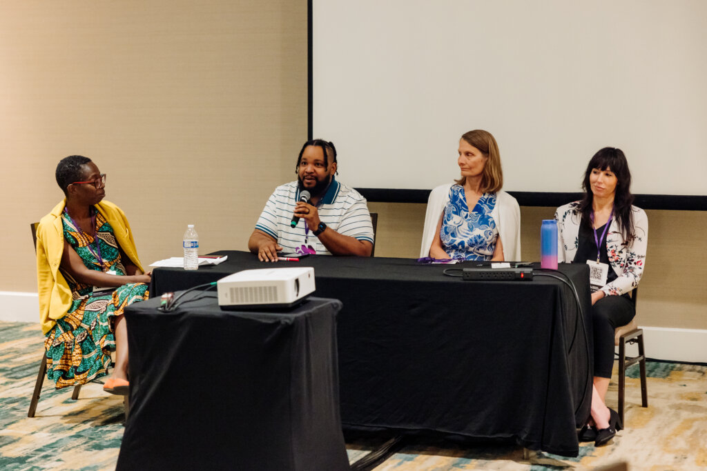 Four people sit at a panel discussion table; one person speaks into a microphone while the others listen. There is a projector and a water bottle on the table, and a blank projection screen behind them.