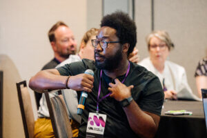 A man with glasses and a beard speaks into a microphone during a conference, wearing a lanyard and badge. Other attendees are seated at a table in the background, listening attentively.