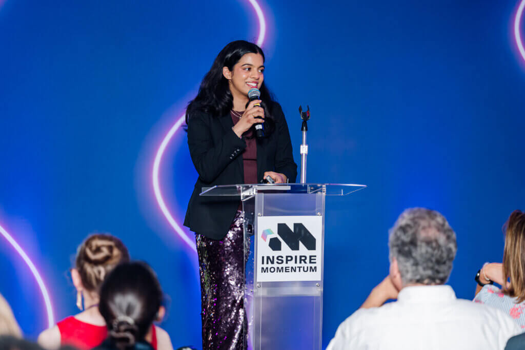A woman stands at a clear podium labeled INSPIRE MOMENTUM, speaking into a microphone to an audience. She is wearing a black blazer and sparkling purple pants, with a blue background behind her.