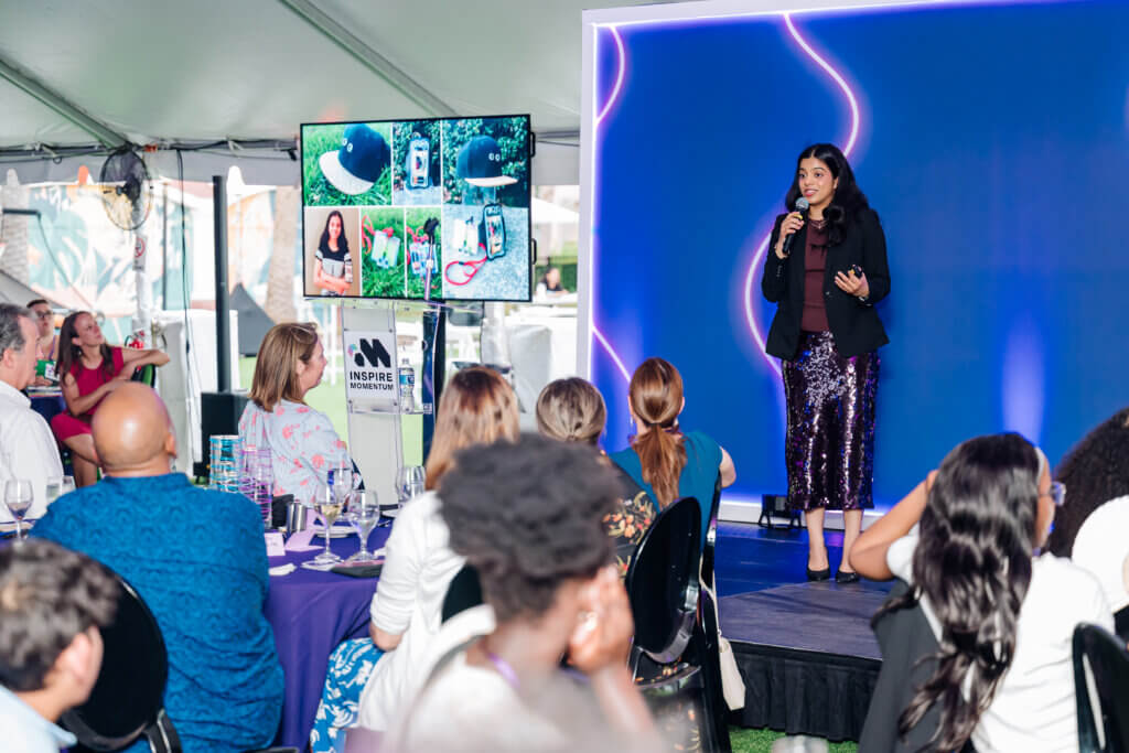 A woman speaks on stage with a microphone in front of an audience at an indoor event. A screen beside her displays photos and graphics. People seated at tables listen attentively.