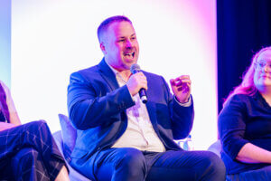 A man in a suit passionately speaks into a microphone while seated on a panel, gesturing with his hand. Two other people sit beside him on stage under vibrant, colorful lighting.
