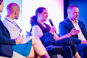 Four people sit on a panel at an event. A woman in the center holds a microphone, speaking while others listen. They are dressed in business attire, with colorful lighting in the background.