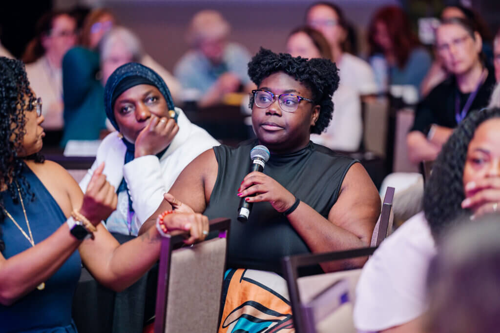 A woman holding a microphone speaks during a group discussion at an indoor event, surrounded by other attentive participants sitting at tables.