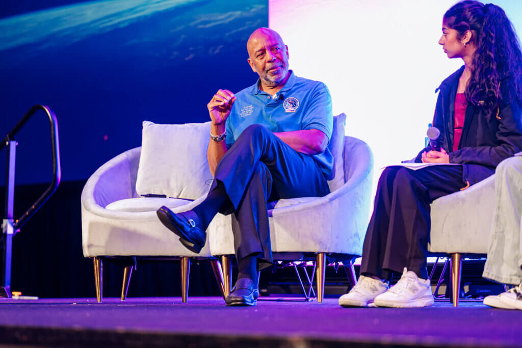 A man in a blue shirt sits on a white couch, speaking and gesturing, while a woman in dark clothing with a notepad listens beside him on stage. The background is blue and purple, and they appear to be at an event or panel.