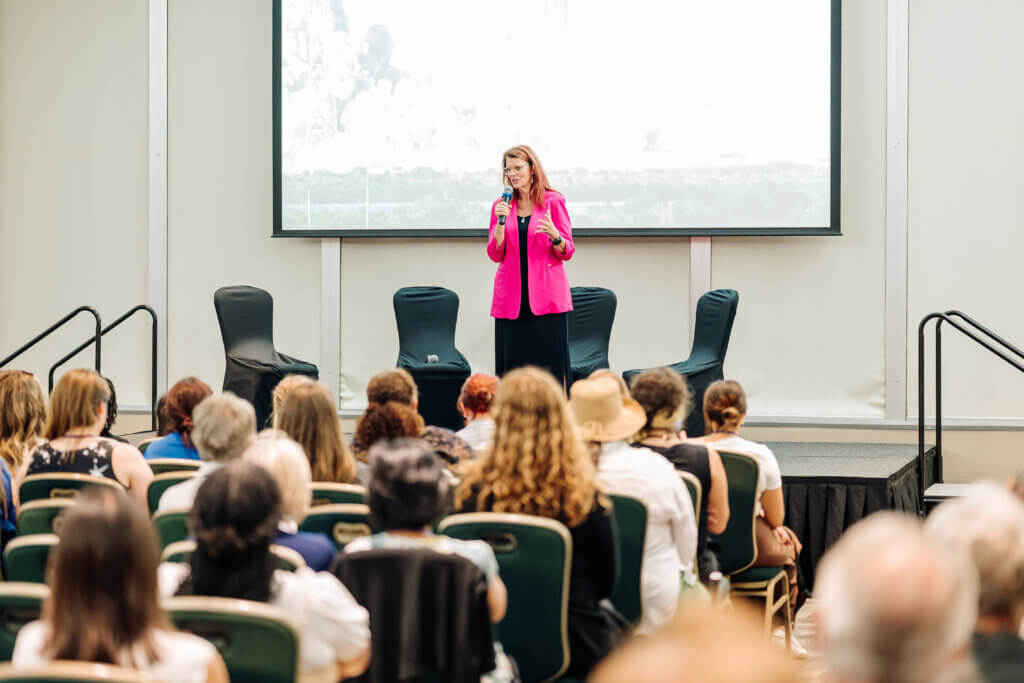 A woman in a pink blazer speaks into a microphone on stage in front of a seated audience in a conference room, with a large screen behind her and two empty chairs on stage.