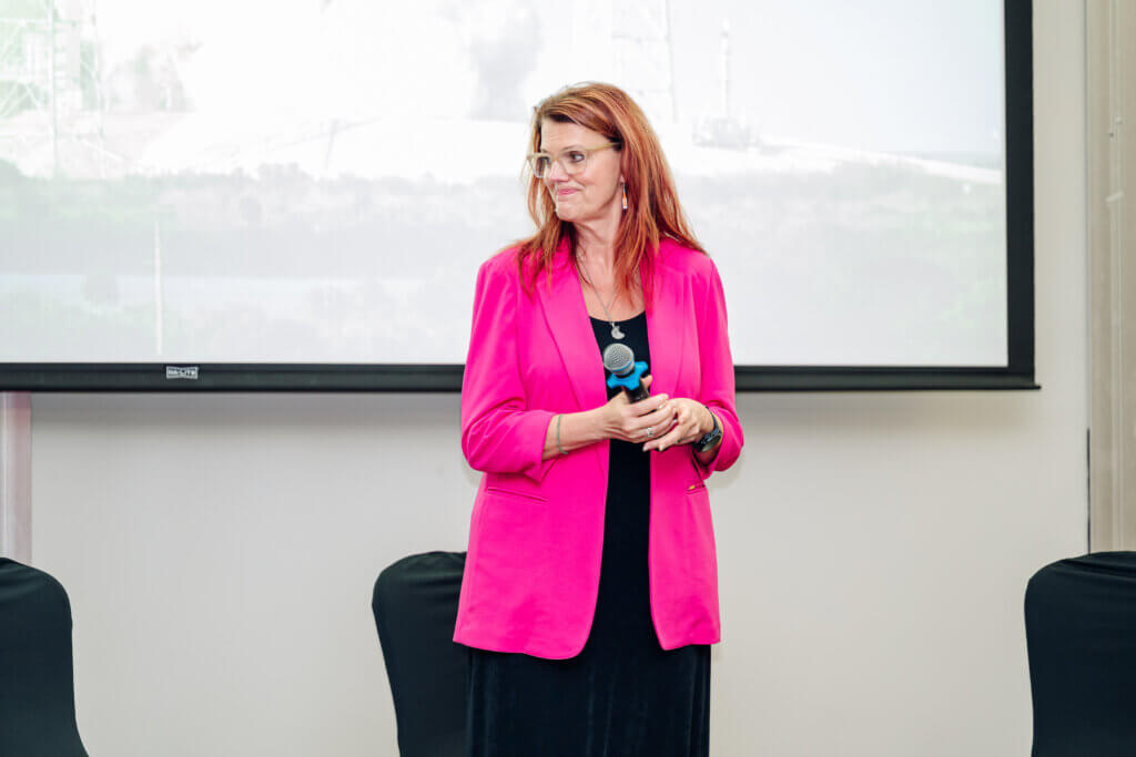 A woman with long red hair, wearing glasses and a bright pink blazer, stands holding a microphone in front of a projection screen in a conference room.