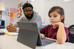 A young girl sits at a table using a tablet with a keyboard, exploring AI literacy, while an adult man sits beside her, watching attentively. Classroom drawings are visible on the wall behind them.