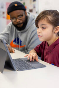 A young girl uses a tablet with a keyboard at a desk, while an adult wearing glasses and a beanie sits nearby, watching and offering support.