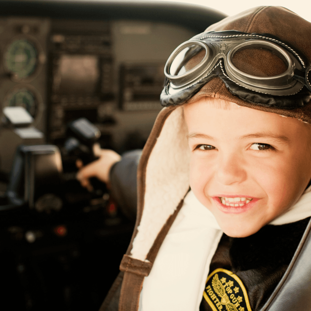 Smiling child in an aviator jacket and goggles sits in an airplane cockpit, holding the controls, with flight instruments visible in the background.