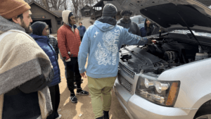 A group of people stand around a car with its hood open outdoors, while one person points inside the engine compartment. Cabins and trees are visible in the background.