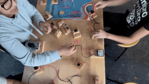 Two people sit at a table assembling simple electric circuits with wires, batteries, and small wooden boards. Various components are spread out on the tabletop.