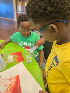 Two children wearing safety glasses conduct a science experiment at a table, pouring liquid into a clear cup. One child wears a green shirt and the other a yellow shirt. Science materials are spread out on the green tablecloth.