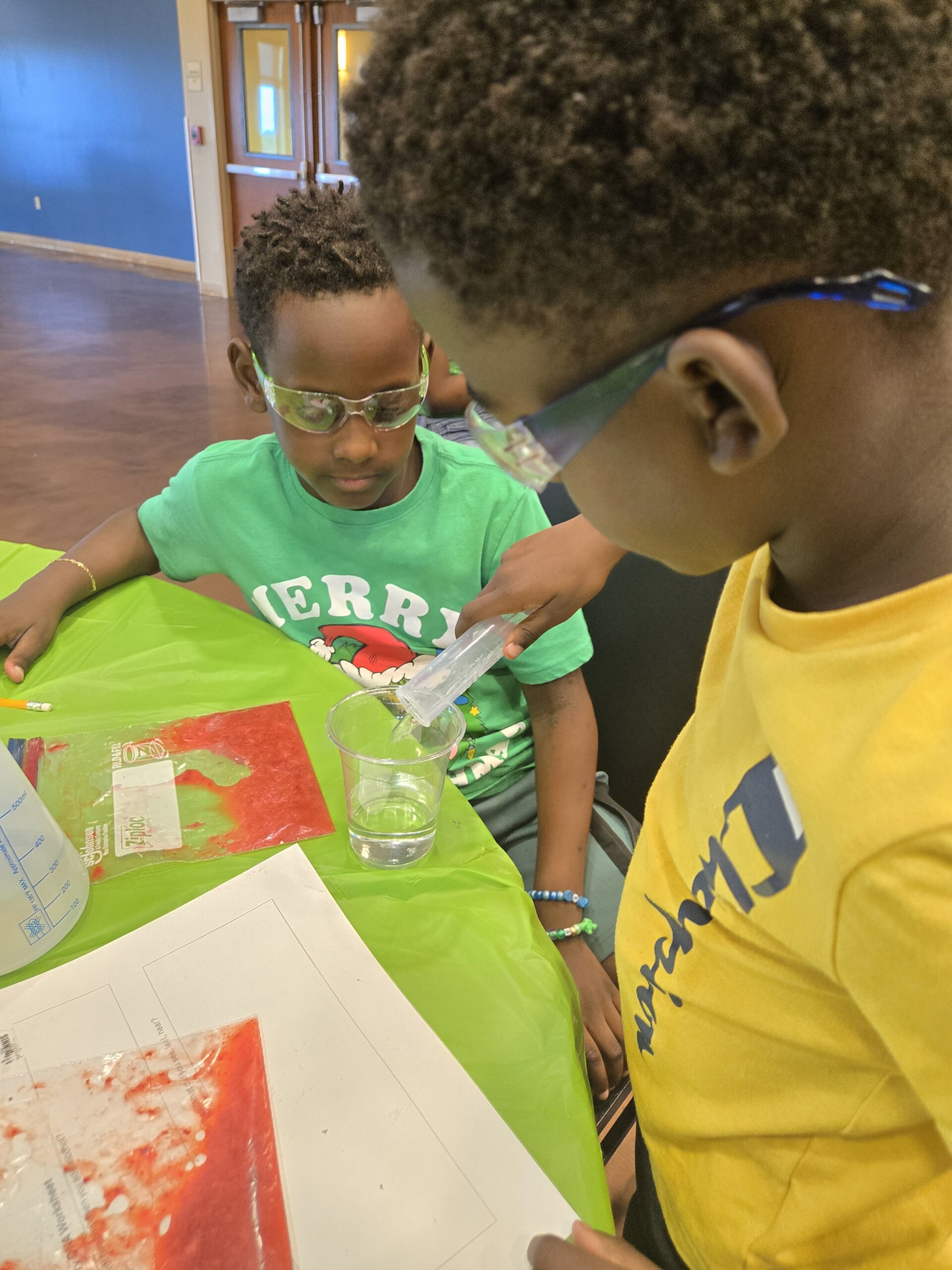 Two children wearing safety glasses conduct a science experiment at a table, pouring liquid into a clear cup. One child wears a green shirt and the other a yellow shirt. Science materials are spread out on the green tablecloth.