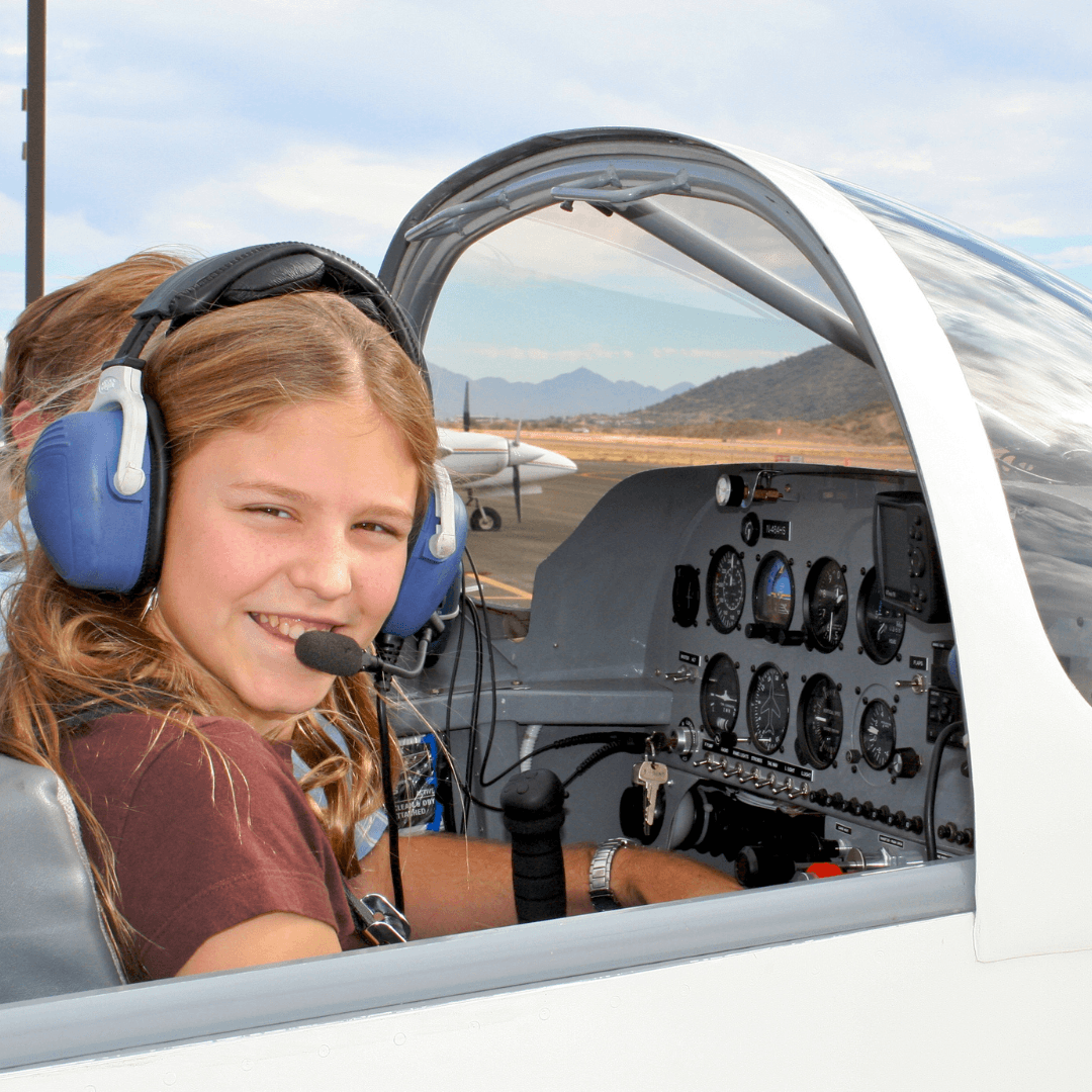 A girl wearing a headset and brown shirt sits in the cockpit of a small airplane, smiling at the camera. Airplane instruments and controls are visible, and there is a runway and mountains in the background.