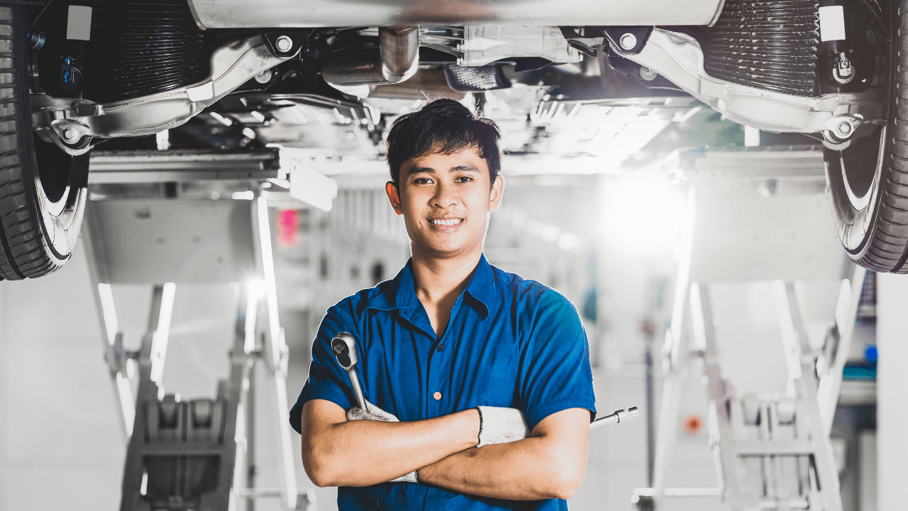 A mechanic in a blue uniform stands smiling with arms crossed, holding a wrench, beneath a car lifted on a hydraulic lift in a brightly lit auto repair shop.