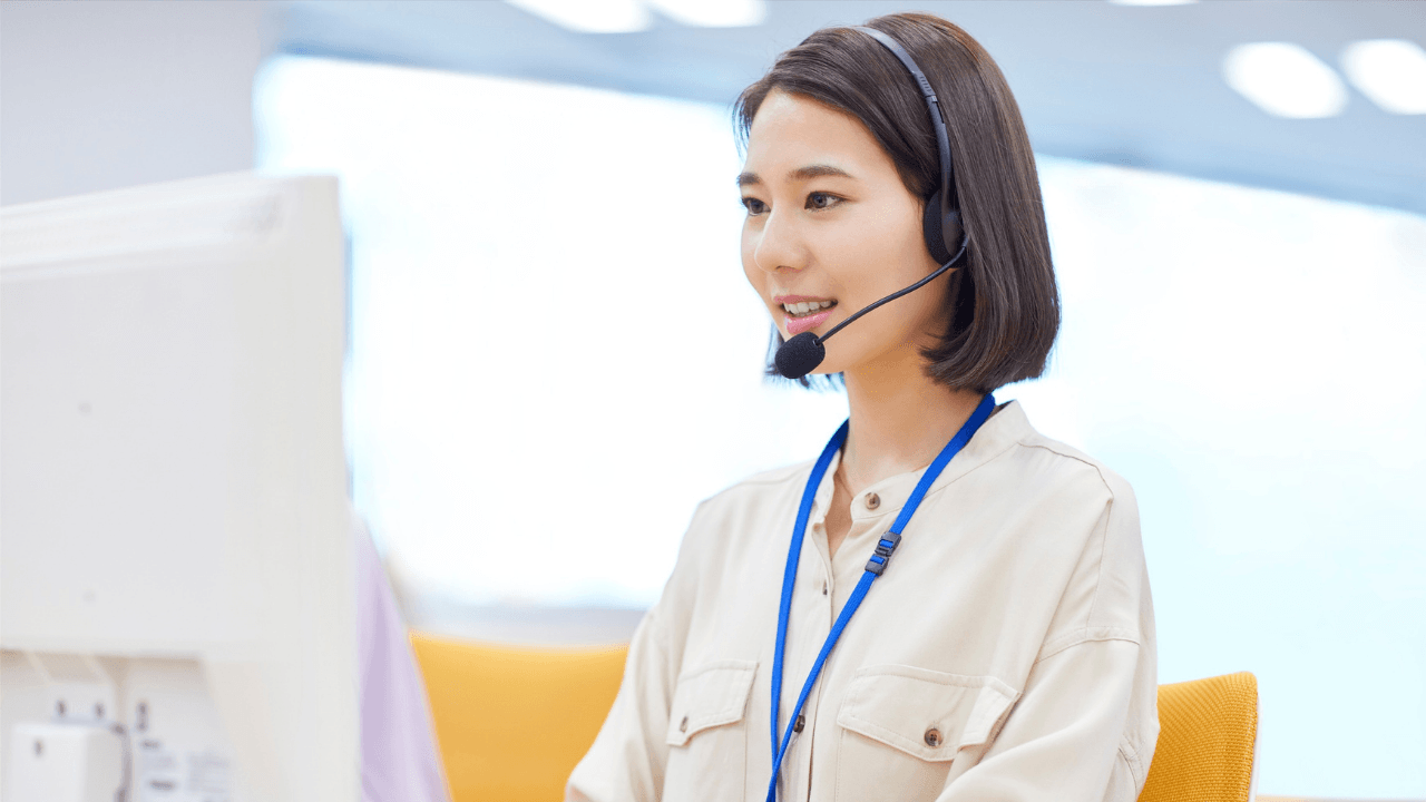 A woman wearing a headset and a lanyard sits at a desk, smiling while looking at a computer monitor in a brightly lit office.