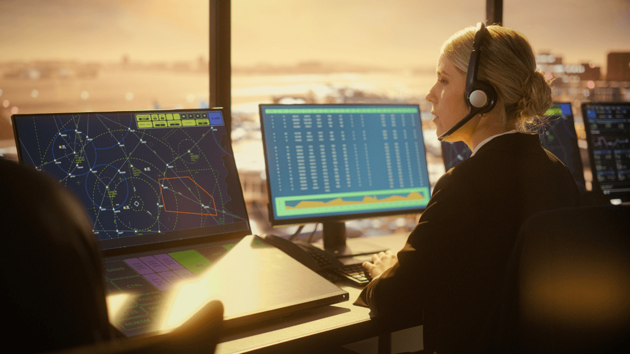 A female air traffic controller wearing a headset works at a desk in a tower overlooking an airport at sunset, monitoring multiple computer screens displaying flight paths, radar data, and technical flight information.
