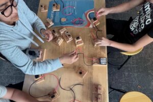 Three children sit around a table assembling circuits with wires, batteries, and wooden blocks. The table is scattered with components and a booklet. One child is seated on a yellow stool.