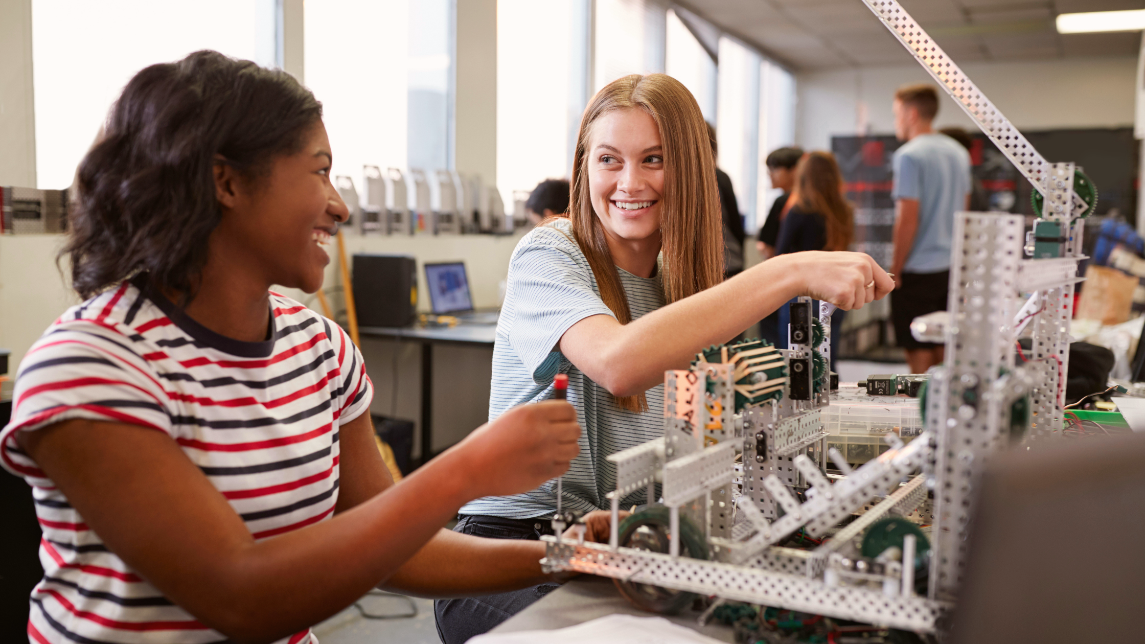 Two young women smile while assembling a metal robotics project in a classroom setting, with computers and other students working in the background.