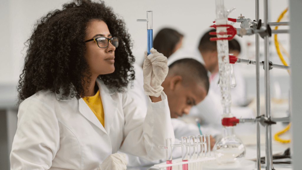 A scientist wearing a white lab coat and gloves examines a test tube with blue liquid in a laboratory. Several glassware and equipment are on the table, and another person works in the background.