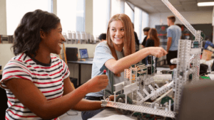 Two young women smile and work together on a robotics project in a classroom, surrounded by laptops and metal robot parts, with other students in the background.