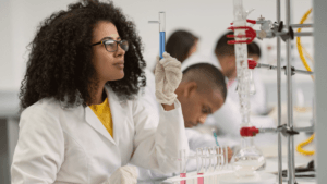 A scientist wearing a white lab coat and gloves examines a test tube with blue liquid in a laboratory. Several glassware and equipment are on the table, and another person works in the background.