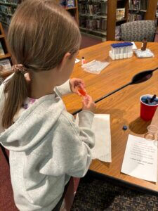 A girl in a light hoodie uses a dropper at a table with science supplies in a library. Nearby is a paper with instructions and labeled tubes, with bookshelves in the background.