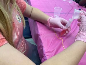 A person wearing pink gloves and a pink apron pours a red liquid into a test tube using a funnel at a table covered with a pink tablecloth.