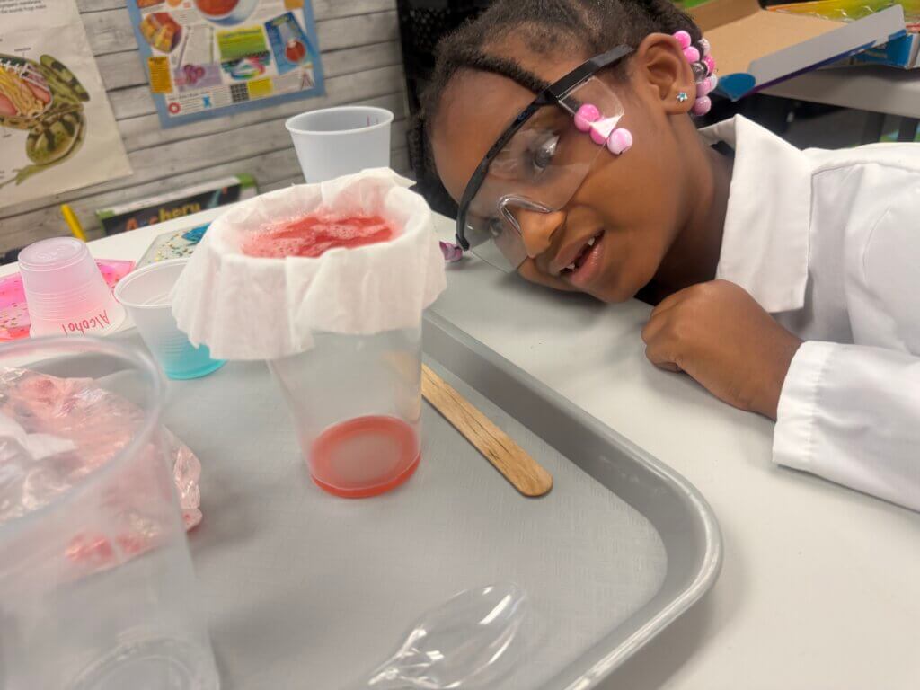 A young girl wearing safety goggles and a lab coat smiles while observing a science experiment with a cup, filter paper, and red liquid on a tray. Science materials are scattered around her on the table.