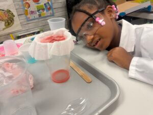 A young girl wearing safety goggles and a lab coat smiles while observing a science experiment with a cup, filter paper, and red liquid on a tray. Science materials are scattered around her on the table.