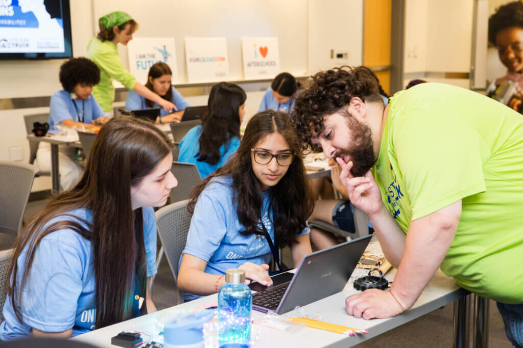 Three people collaborate at a table, two seated with a laptop and one standing, leaning in to help—demonstrating the spirit of AI innovation. Others work in the background, while classroom posters and supplies are visible.
