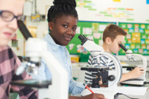 Three students in a classroom sit at desks using microscopes. The student in the center looks toward the camera and smiles while others focus on their work. The background shows colorful charts and bulletin boards.