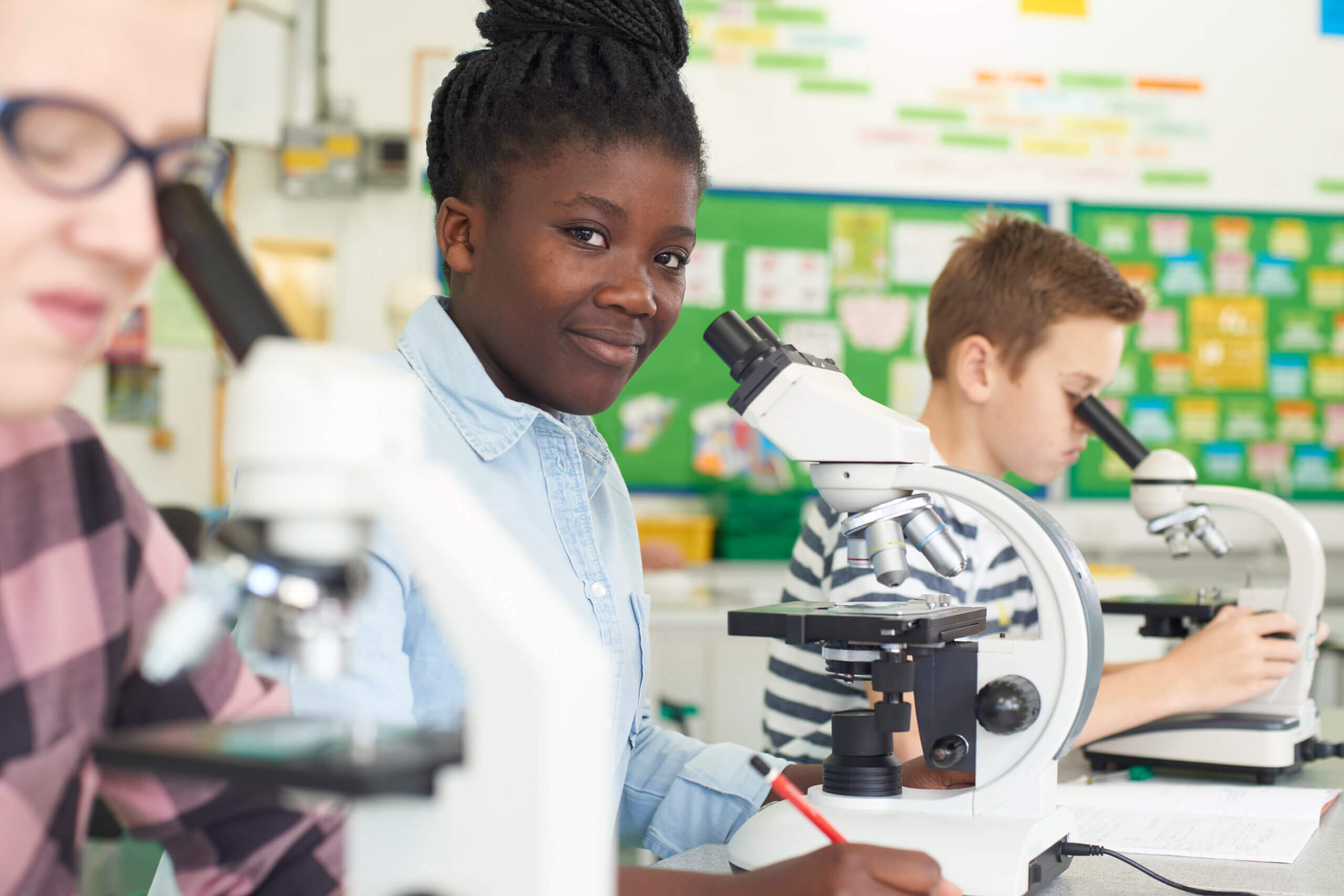 Three students in a classroom sit at desks using microscopes. The student in the center looks toward the camera and smiles while others focus on their work. The background shows colorful charts and bulletin boards.