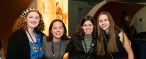 Four young women stand together smiling, wearing conference badges and semi-formal outfits, posing indoors at an event with a warmly lit background. As proud STEM ambassador teens, they represent youth STEM voices making a difference in their community.